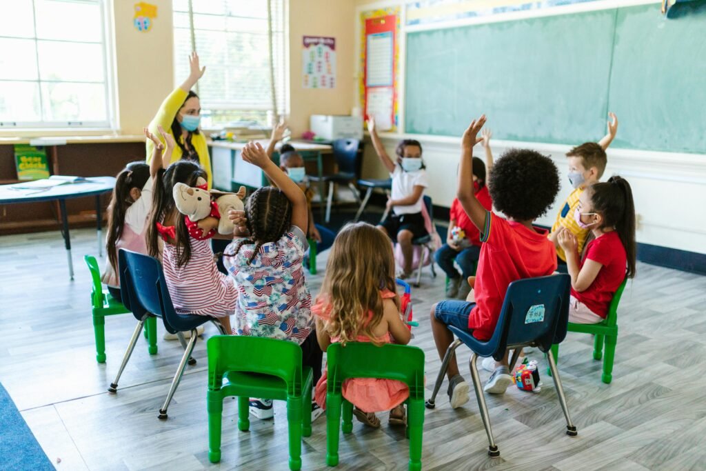 pexels-photo-8364026-8364026 Diverse group of children in a classroom, raising hands, led by masked teacher. Bright and engaging learning environment.